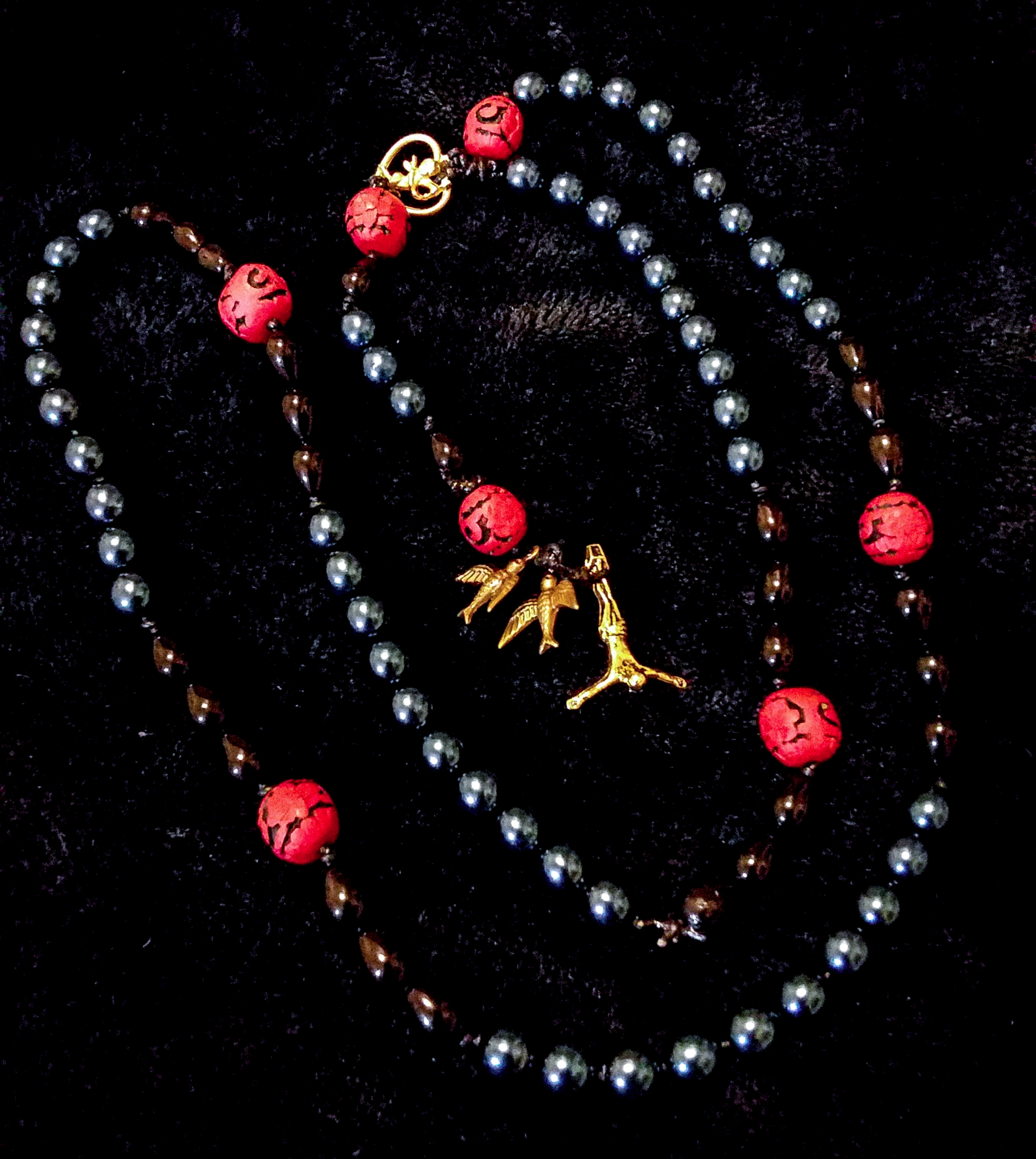 my judas rosary. at the end is an inverted crucifix paired with two sparrow shaped beads. the 'hail holy queen' bead is in the shape of cupid inside a heart. the 'hail mary' beads are all blue pearls, and the 'our father' beads are red with flowers carved into them. these are spaced out with additional black teardrop shaped beads.